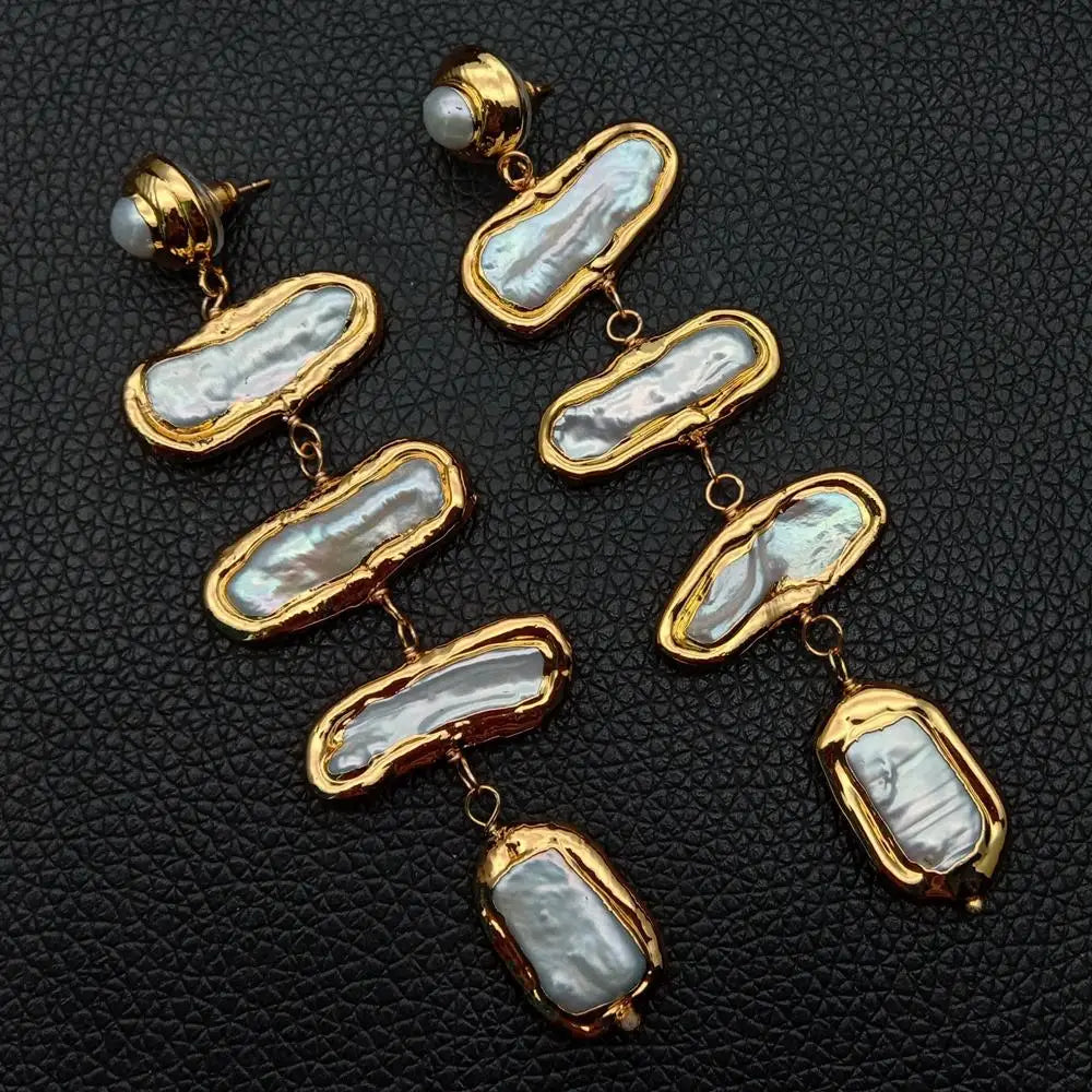 A pair of gold-plated dangle earrings featuring irregular baroque mother-of-pearl inlays, displayed on a textured black leather background.