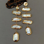 A pair of gold-plated dangle earrings featuring irregular, baroque-style white pearl stones in bezel settings, displayed on a textured gray surface next to a black branded card.