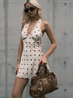 Woman in a polka dot dress holding a brown handbag against a concrete wall.