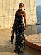 Woman in a black polka dot dress standing by a wooden wall with a scenic background.