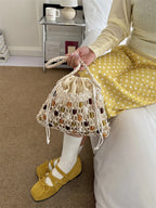 A person seated on a bed holds a cream crochet drawstring bag with woven multicolor accents. The person wears a pale yellow long-sleeve top, a yellow skirt with white polka dots, white socks, and mustard yellow flats in a bedroom setting.