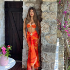 Woman in an orange outfit standing in a stone doorway with flowers and a mirror in the foreground.