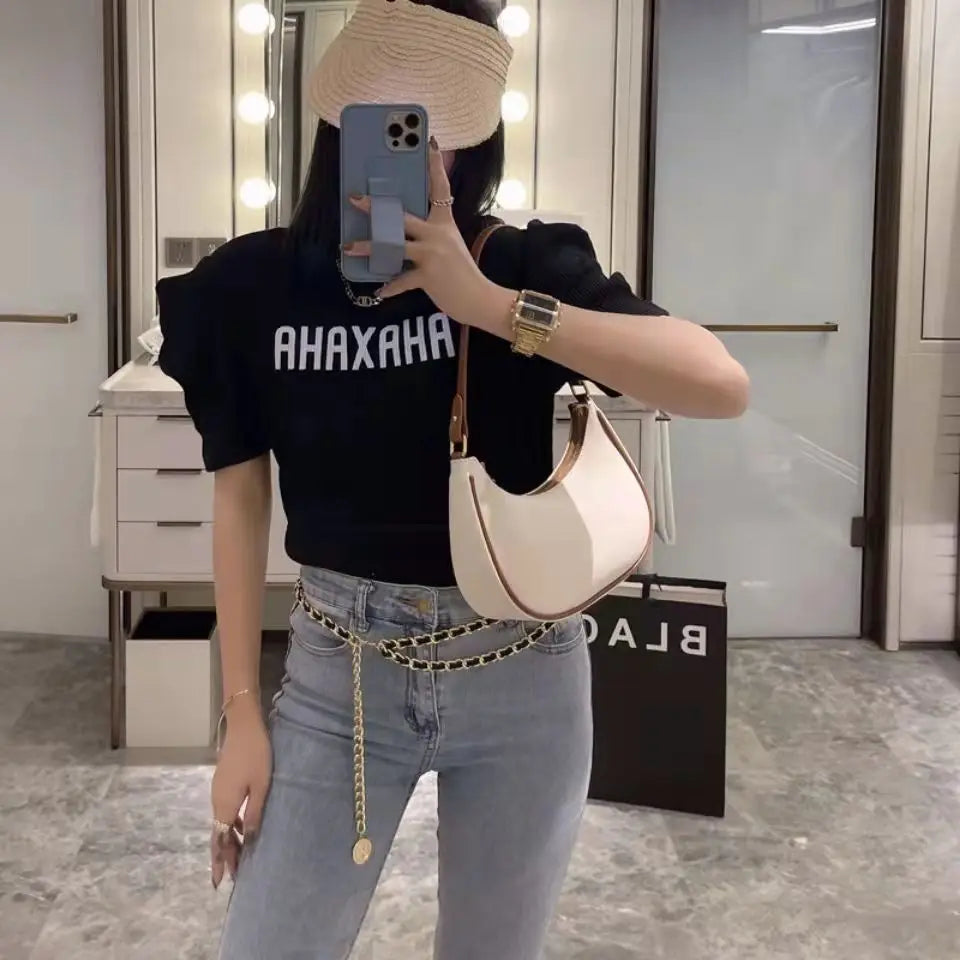 Person wearing a black t-shirt with 'AHAXAHA' text, light blue jeans, and a beige handbag in a mirror selfie.