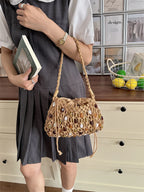 A person wearing a gray pleated dress and white short-sleeve blouse holds a woven straw shoulder bag with a braided strap in front of a cream-colored dresser and framed wall art.