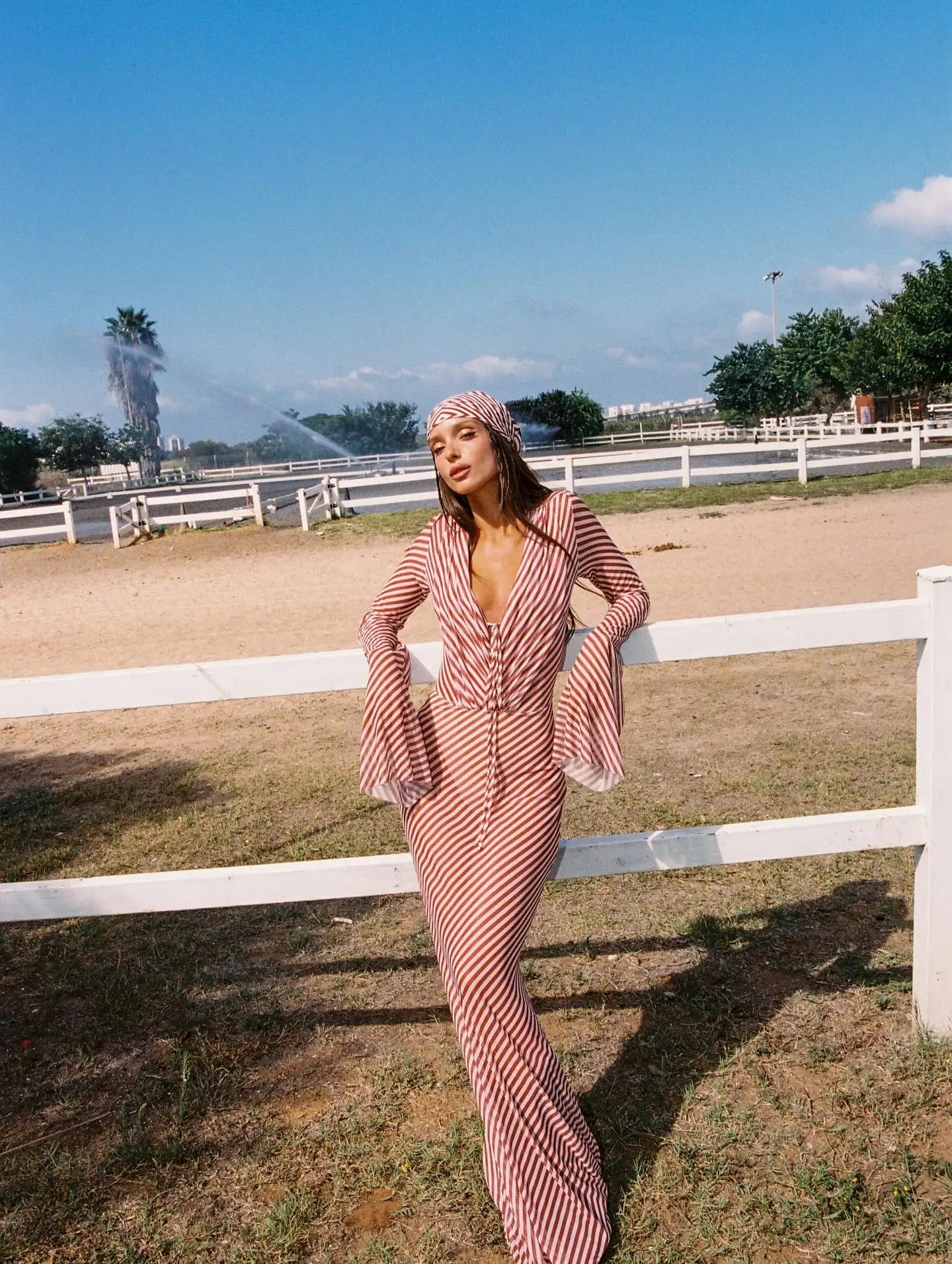 Woman in a pink dress standing in an outdoor setting with a white fence and trees in the background.