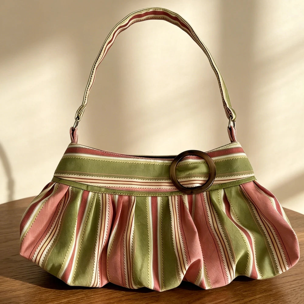 A pleated shoulder bag with vertical stripes in sage green, dusty pink, and cream, featuring a wide matching strap and a prominent round brown metal ring detail on the front, resting on a wooden table.
