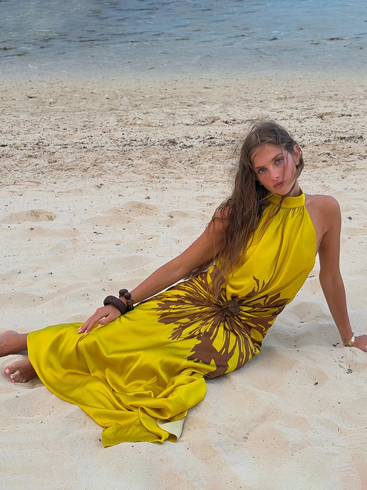 A woman sits on a sandy beach wearing a bright yellow halter maxi dress with a large brown abstract floral print. The background shows the shoreline and water.