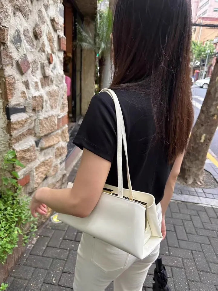 Person holding a white handbag on a street with a stone wall and greenery in the background