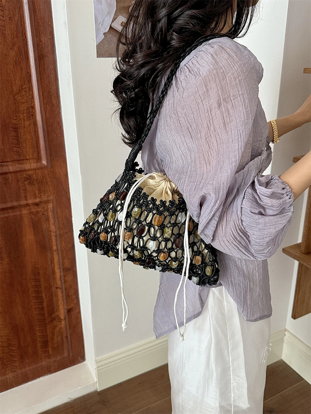 A woman wearing a lavender blouse and white skirt carries a black crochet bucket bag with a drawstring closure and a beige fabric lining. The photo is taken in an indoor setting with a wooden door on the left.