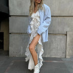 Woman wearing a light blue coat over a white dress with ruffles, standing against a concrete wall.