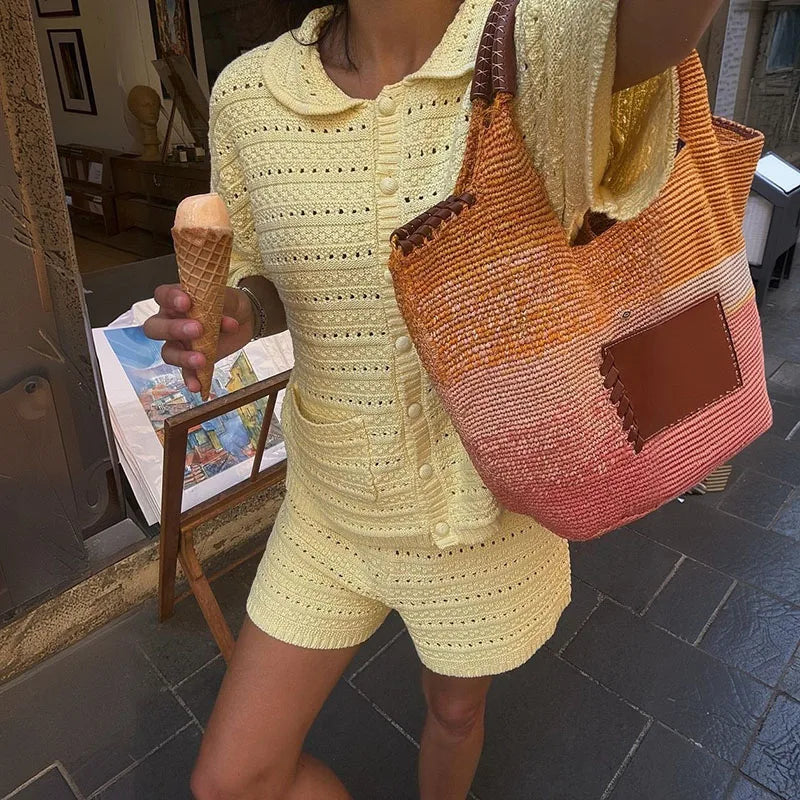 A person wearing a pale yellow crocheted short-sleeve button-front top and matching shorts, holding an ice cream cone and carrying a large woven tote bag with orange, yellow, and pink tones in an outdoor setting.