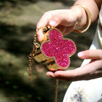 Hand holding a pink glittery clover-shaped bag with gold accents against a blurred natural background.