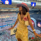 A woman wearing a yellow floral mini dress with a red hat and a watch, standing in a stadium with a crowd in the background.