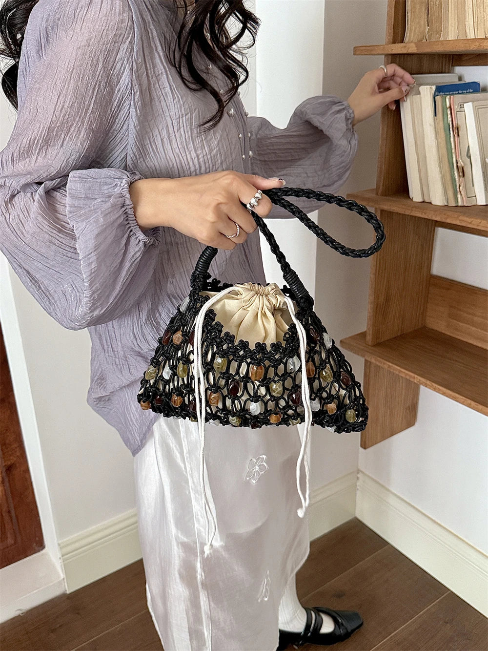 A person in a light lavender blouse and white skirt holds a black crochet bucket bag with a cream drawstring and multicolored stone-like accents, standing indoors near a wooden bookshelf.