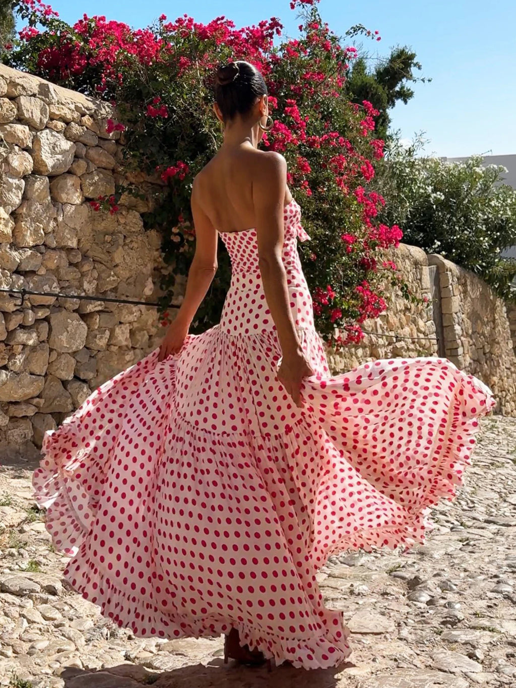 Woman in a red and white polka dot dress standing in front of a stone wall and flowering bush.