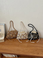 Three crocheted bucket bags with woven stone accents are lined up on a wooden shelf against a textured off-white wall. The bags are in natural tan, cream, and black, each with a braided shoulder strap and drawstring closure.