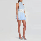 A model wearing a light blue halter mini dress with a white lace waist detail, paired with silver high-heeled sandals, against a light gray studio background.