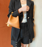 Person wearing a black blazer and shorts holding a tan handbag against a textured wall.