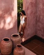 Woman in a white swimsuit standing near rustic vases against a textured wall.