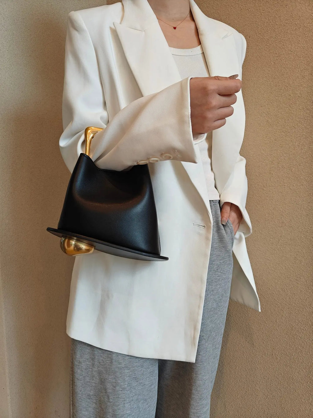Person wearing a white blazer and gray pants holding a black handbag with gold accents against a beige background