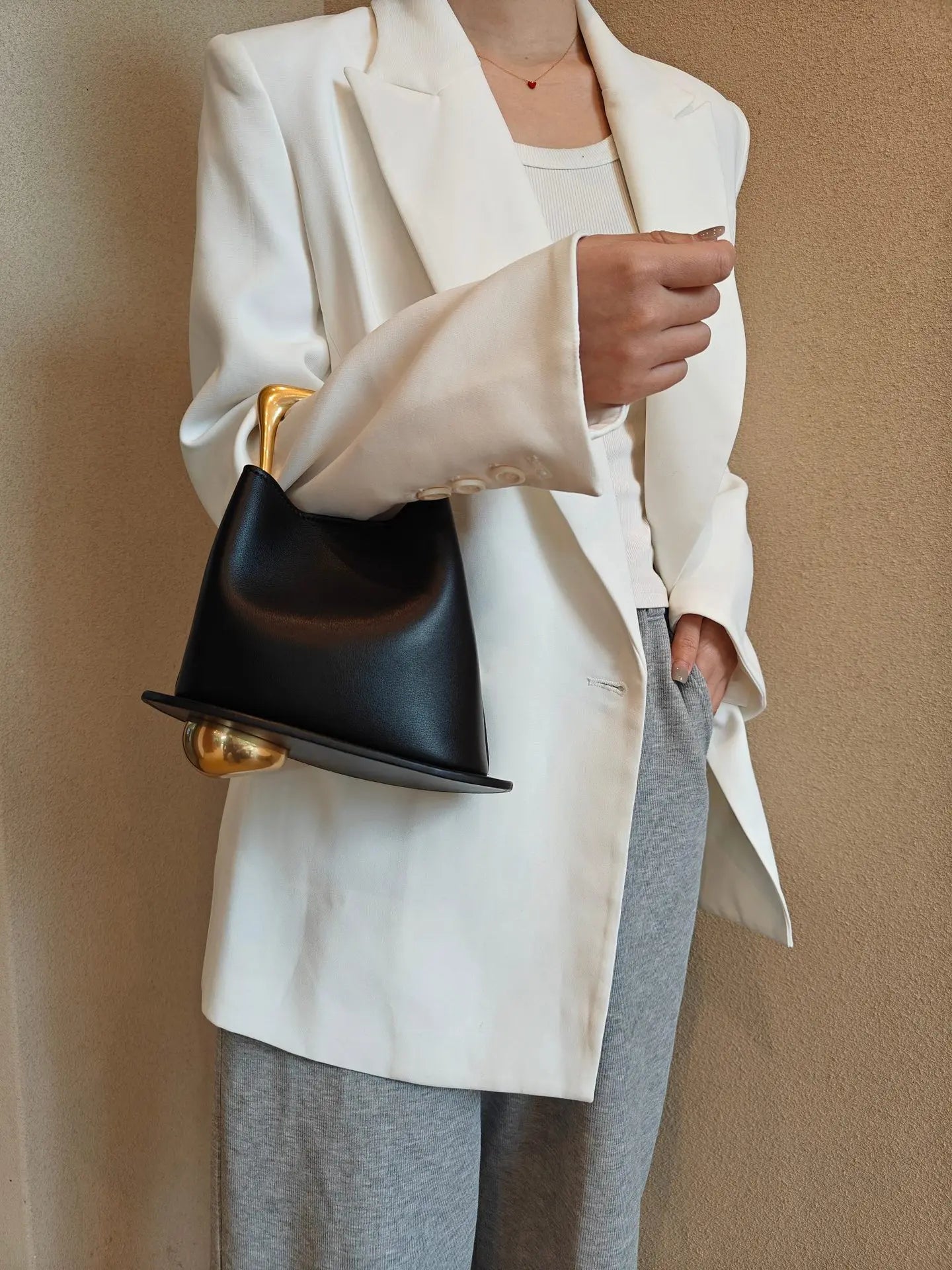 Person wearing a white blazer and gray pants holding a black handbag with gold accents against a beige background