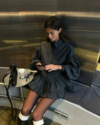 A woman sits on a bench in a modern interior with metallic walls, wearing a dark gray oversized collared top with voluminous sleeves and a matching ruffled mini skirt. She is looking down at a phone, with a black handbag, a coffee cup, and glasses on a small side table beside her.