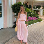 A woman stands outdoors on a paved walkway wearing a light pink two-piece outfit with a lace-trimmed crop top and matching loose harem-style pants. She is holding a woven straw bag, with a pink building, green plants, and pink flowers in the background.