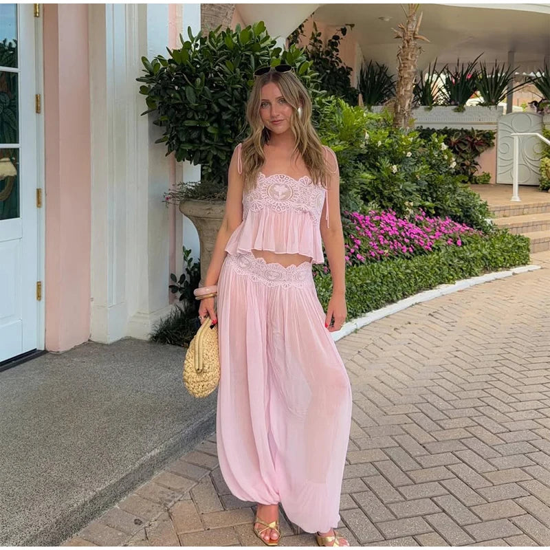 A woman stands outdoors on a paved walkway wearing a light pink two-piece outfit with a lace-trimmed crop top and matching loose harem-style pants. She is holding a woven straw bag, with a pink building, green plants, and pink flowers in the background.