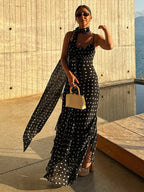 Woman in a black polka dot dress with a scarf and sunglasses, standing by a waterfront.