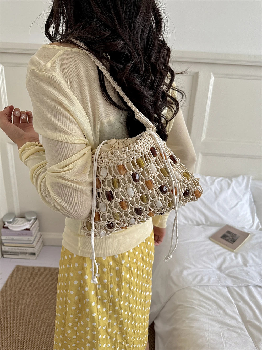A woman wearing a cream knit long-sleeve top and a yellow polka-dot skirt carries a cream macrame shoulder bag with woven brown and amber stones, standing in a bedroom setting.