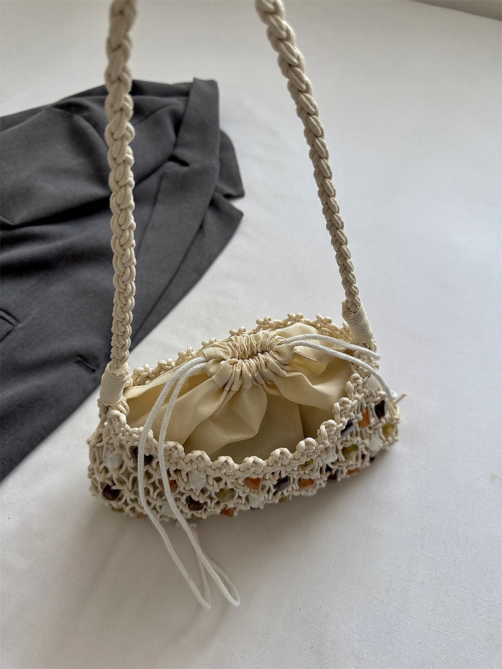 A cream macrame shoulder bag with a drawstring pouch, resting on a white surface next to a dark gray fabric prop.