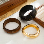 Three bracelets in black, brown, and gold on a textured surface with a wooden box in the background.