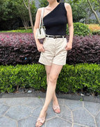 Person wearing a black top and beige shorts standing outdoors with greenery in the background