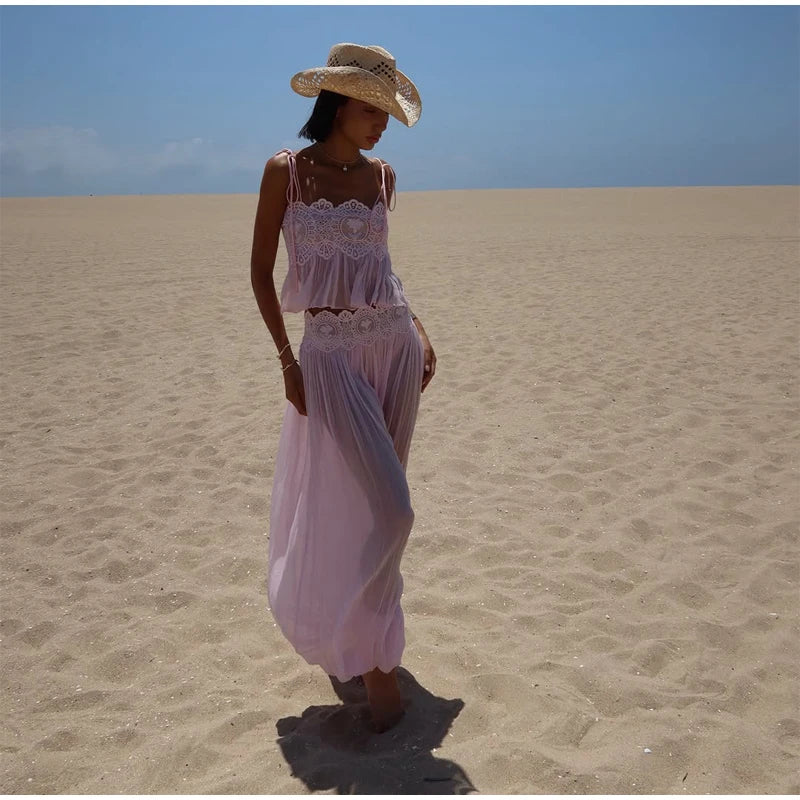 A woman wearing a light pink two-piece dress and a straw hat stands barefoot on a sandy beach under a clear blue sky.