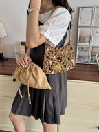 A woman wearing a white short-sleeve top and a dark gray pleated mini skirt carries a chunky woven tan shoulder bag made of circular shell-like elements, while also holding a tan drawstring pouch in front of a home interior background.