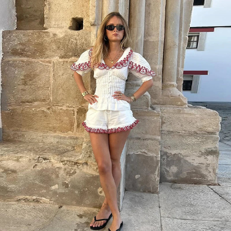 Woman in a white outfit with red embroidery standing against a stone wall.