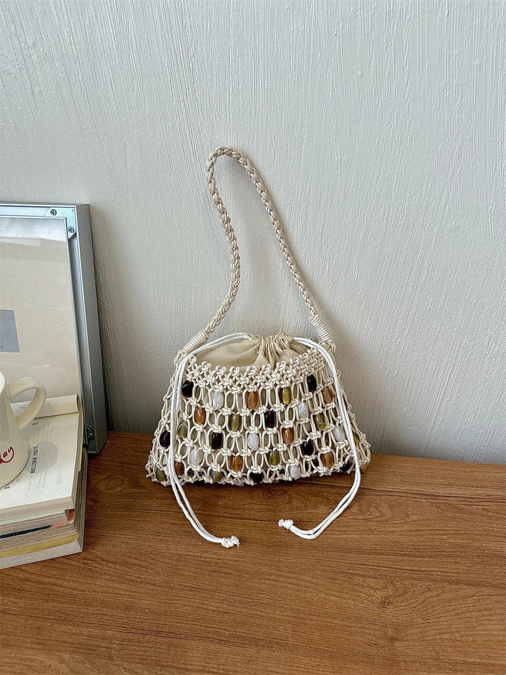 A cream macrame drawstring bucket bag with brown and gold woven accents, resting on a wooden table against a textured light gray wall.