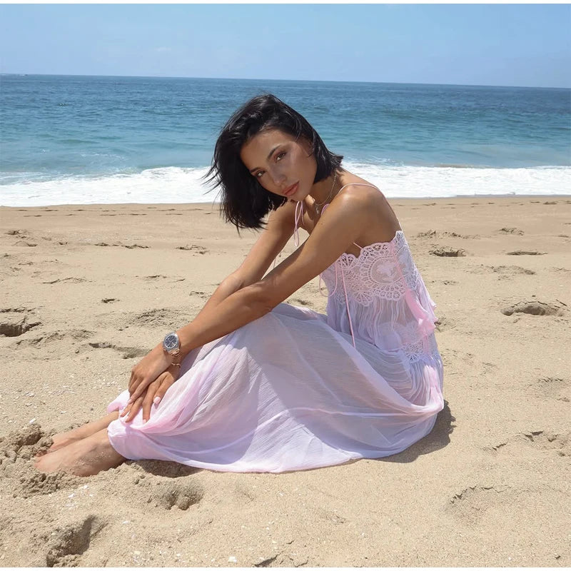 A woman sits on a sandy beach wearing a sheer light pink dress with a white lace bodice, with the ocean and blue sky in the background.