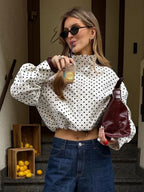 Woman in polka dot blouse and sunglasses holding a drink and a bottle, standing on steps with fruit crates in the background.