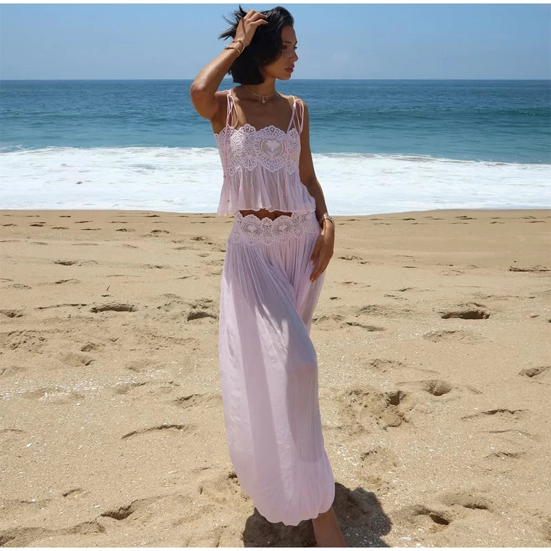 A woman stands on a sandy beach wearing a light pink two-piece outfit with a lace-trimmed tank top and matching maxi skirt, with the ocean and blue sky behind her.