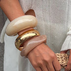 Close-up of a hand wearing two large bangles, one gold and one beige, against a neutral background.