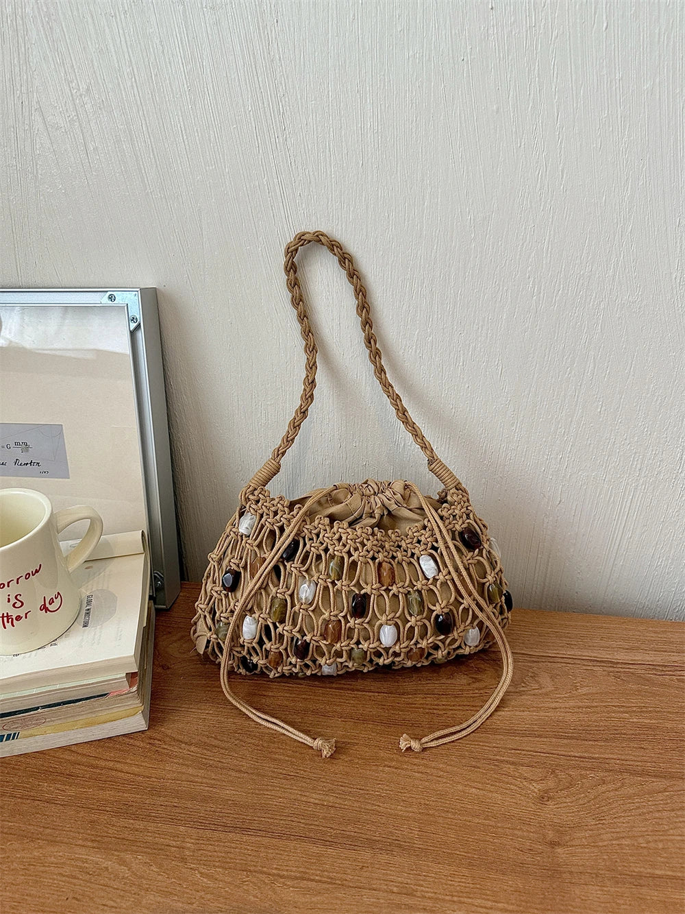 A beige woven macrame-style drawstring handbag with shell embellishments sits on a wooden table next to a stack of books and a white ceramic mug against a textured off-white wall.