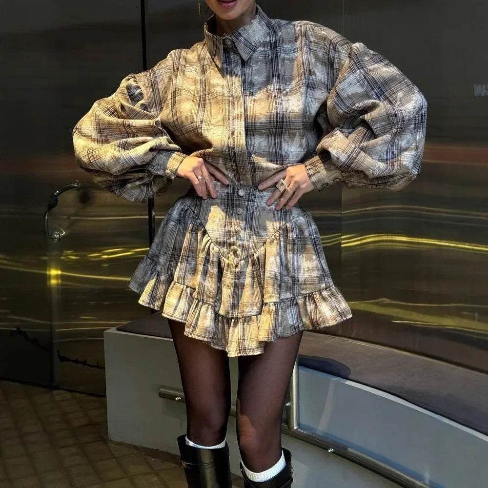 A person wearing a beige and brown plaid mini shirt dress with voluminous balloon sleeves, a high collar, and a ruffled tiered hem, paired with sheer black tights and black boots.