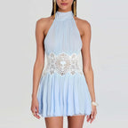 A woman wearing a light blue halter mini dress with a pleated skirt and a white lace embroidered waistband, shown against a plain light gray studio background.