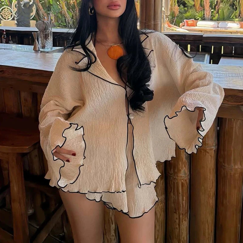 A woman wearing a beige crinkled pajama set with black outline details, featuring a long-sleeve button-up top and matching shorts, posed in front of a rustic wooden bar.