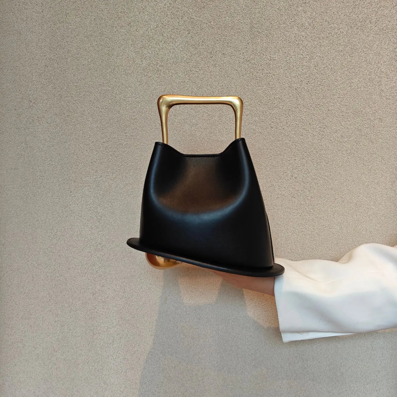 Black handbag with gold handle held by a hand against a beige background