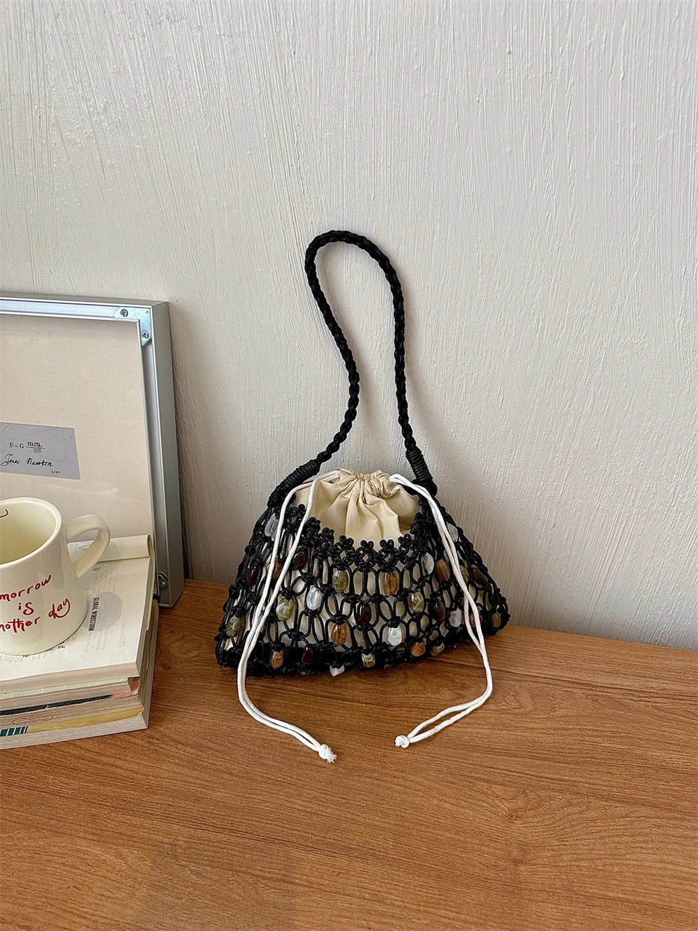 A black crochet bucket bag with a cream drawstring closure and white braided shoulder strap, styled on a wooden table next to a mug and books against a textured wall.
