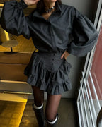 A person wearing a dark charcoal gray mini shirt dress with dramatic puffed long sleeves, a buttoned waist, and layered ruffle details, paired with black tights and black boots in an indoor setting.