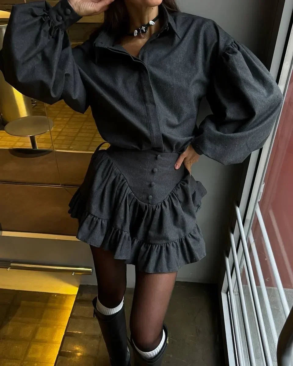 A person wearing a dark charcoal gray mini shirt dress with dramatic puffed long sleeves, a buttoned waist, and layered ruffle details, paired with black tights and black boots in an indoor setting.