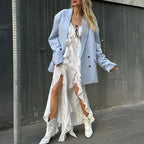 Woman wearing a light blue blazer over a white dress with a high slit, standing against a dark wall.